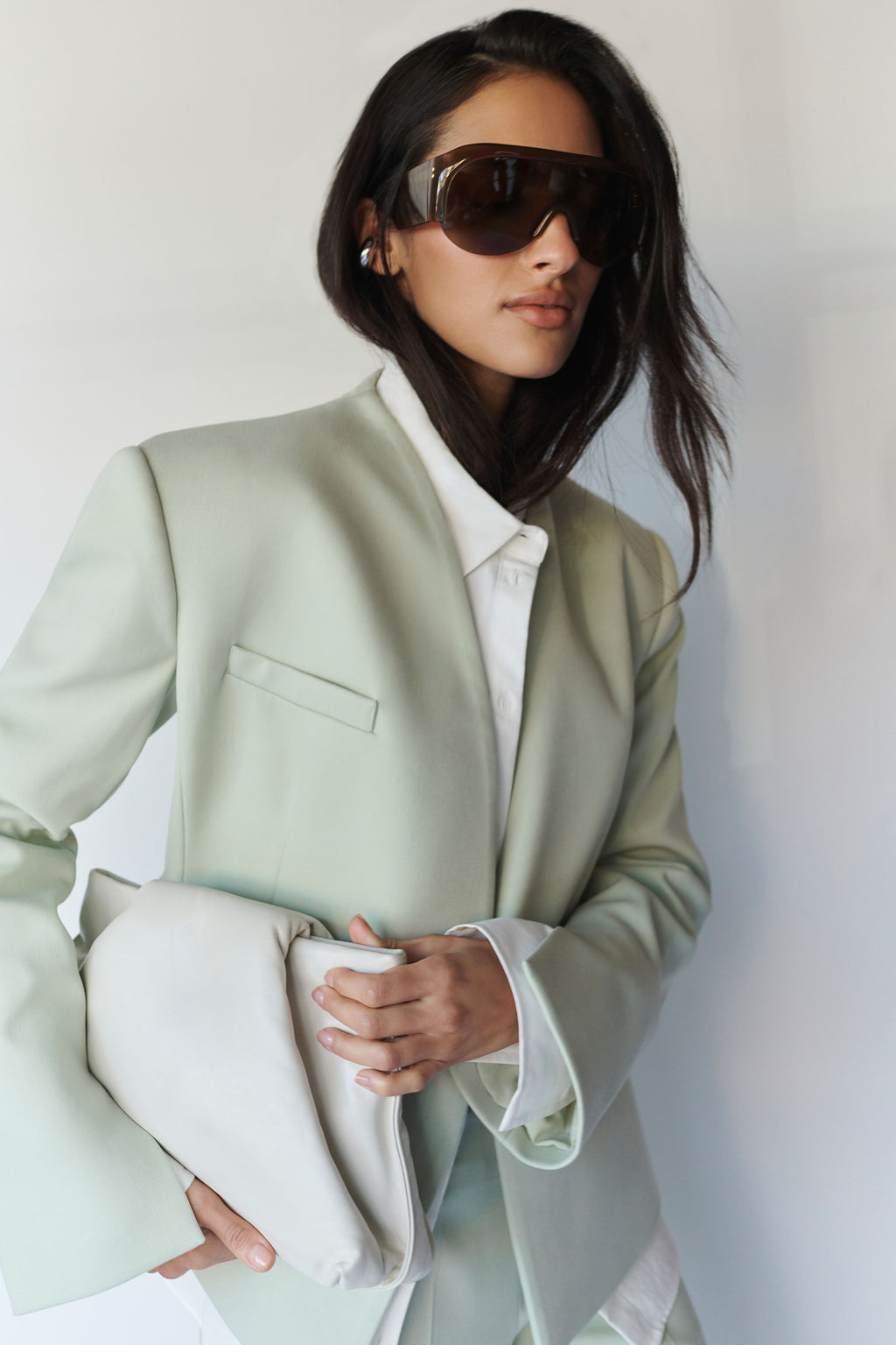 A woman wearing oversized dark sunglasses, a pale sage green blazer over a white collared shirt, and holding a matching sage green clutch bag against a plain light background.