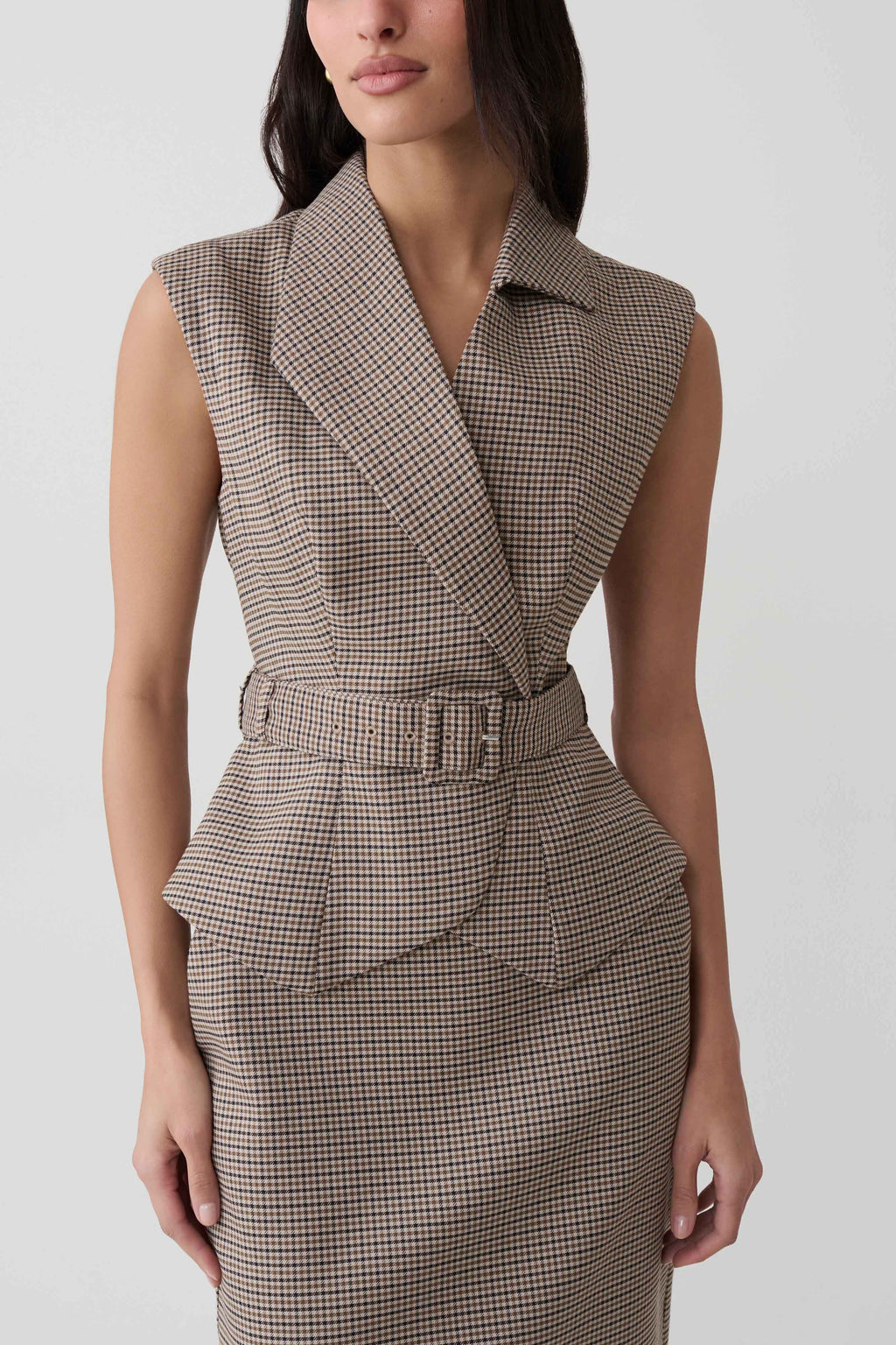 A woman wearing a sleeveless belted wrap dress with a fine brown and beige houndstooth pattern, shown from the neck down against a light gray studio background.