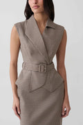 A woman wearing a sleeveless belted wrap dress with a fine brown and beige houndstooth pattern, shown from the neck down against a light gray studio background.