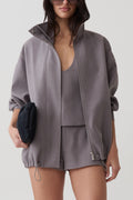 A woman models an oversized gray zip-up jacket with a stand collar, paired with matching shorts and a matching camisole. She holds a small black clutch against a clean, light gray studio background.