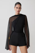 Woman wearing a black outfit with sheer sleeves on a plain background.