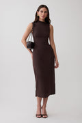A woman modeling a sleeveless, high-neck, knee-length dark brown dress against a plain light gray studio background. She is carrying a small brown shoulder bag and wearing black heeled sandals.