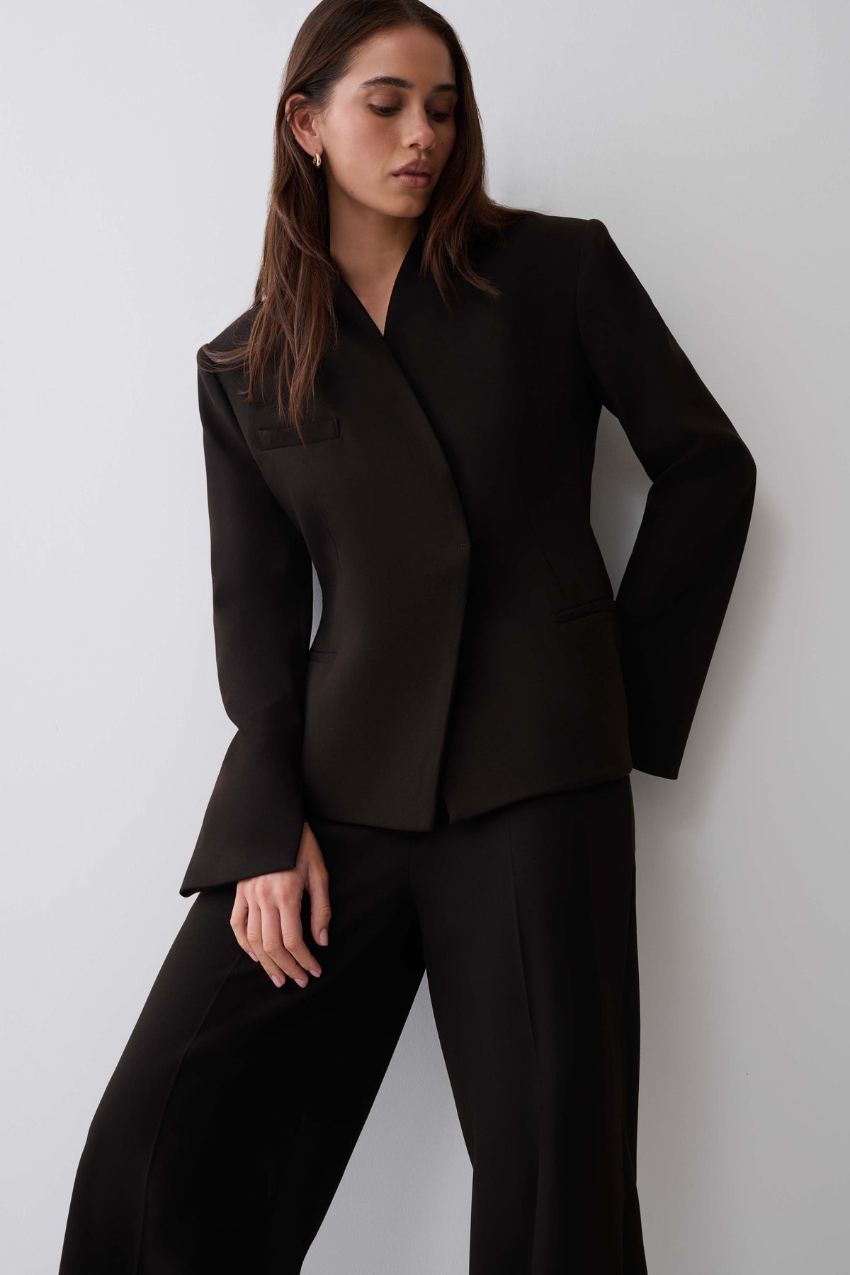 A woman modeling a black tailored blazer with structured shoulders and matching black trousers against a plain light gray studio background.