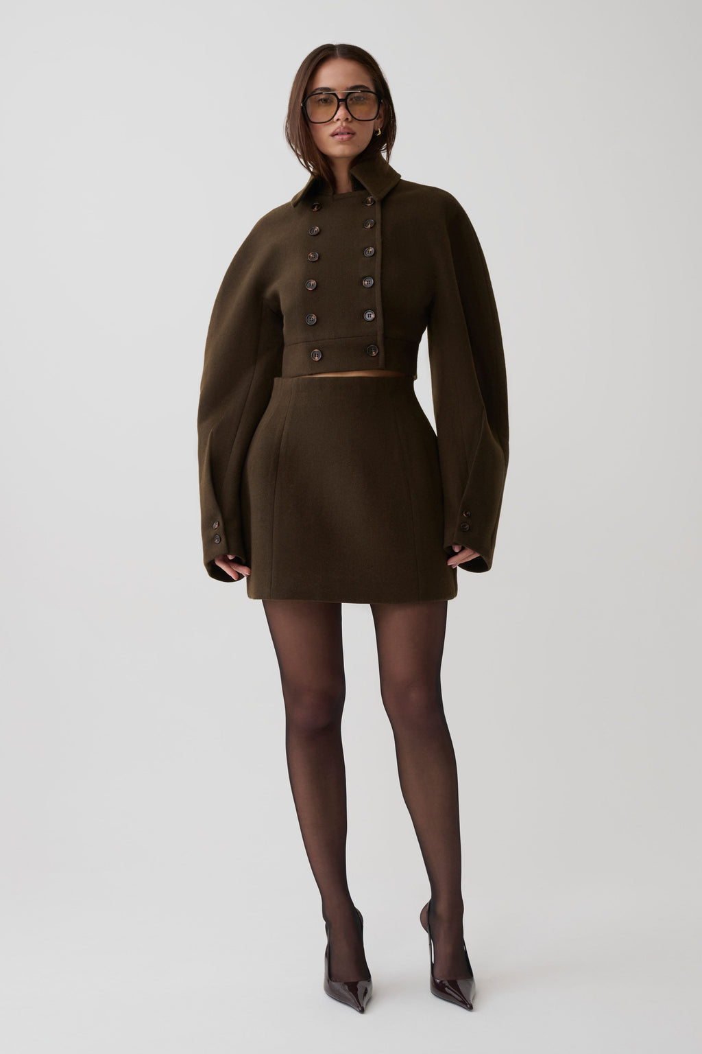 A model wearing a dark brown double-breasted mini coat with oversized sleeves, paired with sheer black tights and dark pointed-toe heels, standing against a light gray studio background.