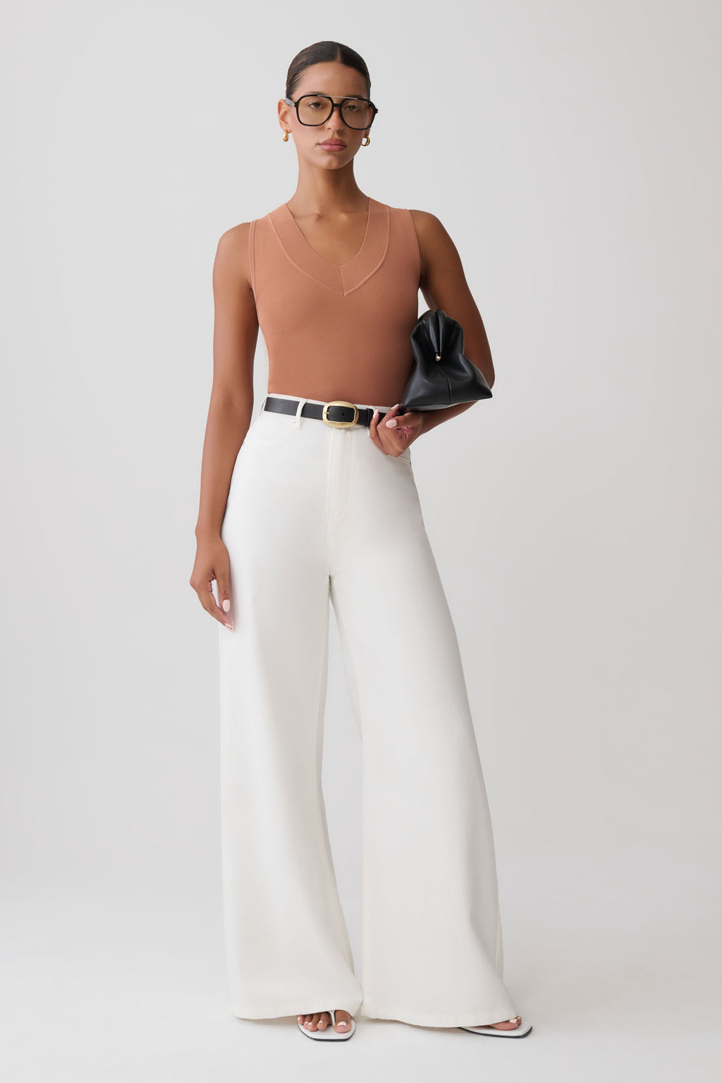 A model stands against a light gray studio background wearing a ribbed tan sleeveless top, high-waisted white wide-leg trousers with a thin black belt, and holding a black clutch.
