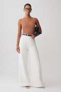 A model stands against a light gray studio background wearing a ribbed tan sleeveless top, high-waisted white wide-leg trousers with a thin black belt, and holding a black clutch.