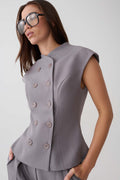 A woman wearing a structured sleeveless peplum-style top in a muted grayish lavender tone, paired with matching trousers and oversized dark sunglasses, against a plain light gray studio background.