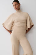 A model wearing a beige knit matching set consisting of a mock-neck top with wide flared sleeves and high-waisted, straight-leg trousers, posed against a light gray studio background.